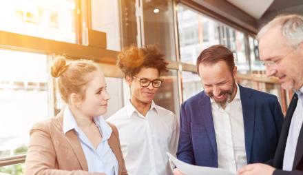 4 Personen im Büro, die freundlich lächeln und auf einem Blatt Papier schauen